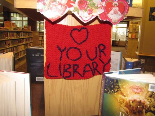 A knitted sign on a library shelf reads "LOVE YOUR LIBRARY" with decorative hearts above it.