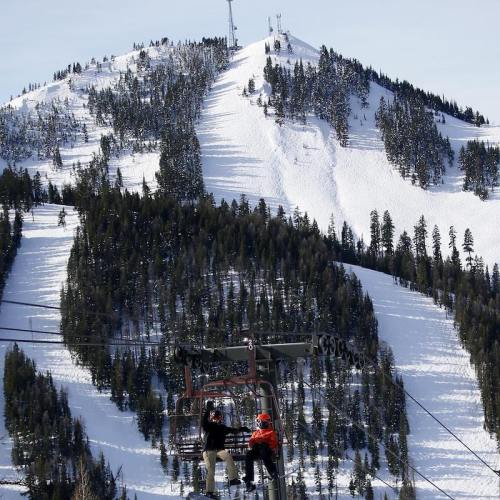 A ski lift carries passengers up a snowy mountain, surrounded by evergreen trees and a clear blue sky.