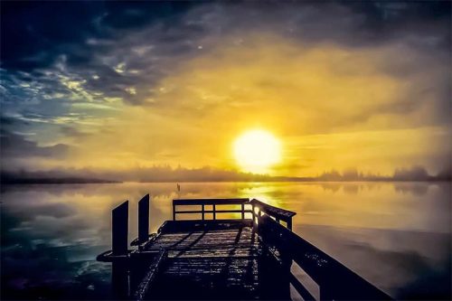 A serene sunrise over a calm lake, viewed from a wooden dock surrounded by mist and soft clouds.
