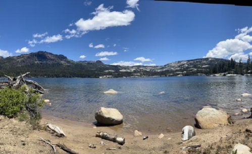 Panoramic view of a serene lake surrounded by mountains and trees, with rocks on the sandy shore under a blue sky.