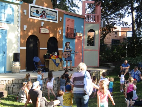 A performer plays guitar on stage for a crowd of children and families in a park setting. Colorful buildings in the background.
