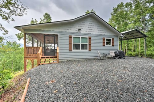 A cozy gray house with a porch, surrounded by trees and gravel, featuring outdoor seating and a grill.