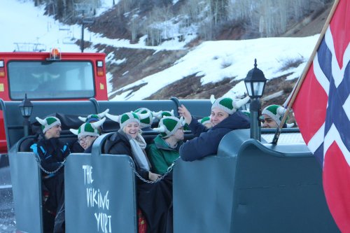 A group of people wearing Viking hats smiles while sitting in a sled, with snowy mountains and a Norwegian flag in the background.