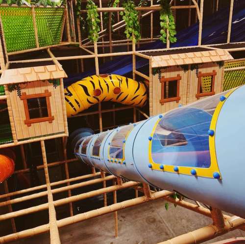 Colorful play structure featuring a slide, tunnels, and treehouse-like structures surrounded by greenery.