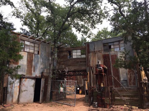 Rustic building made of corrugated metal and wood, surrounded by trees, with a fenced entrance and a weathered appearance.