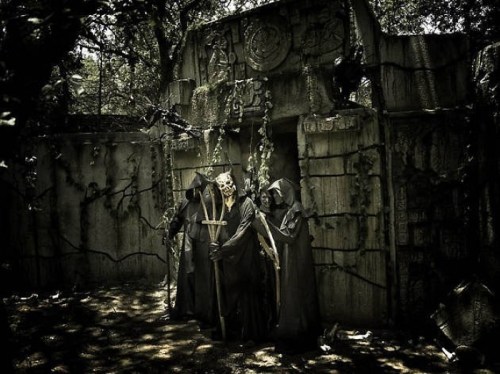 Three cloaked figures stand in front of an ancient, overgrown stone structure, shrouded in darkness and mystery.