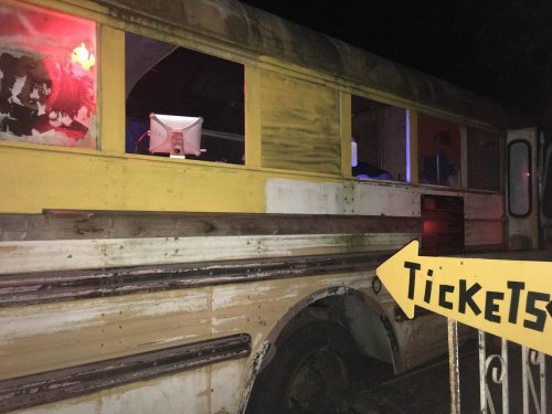 A weathered yellow school bus with a ticket sign, illuminated by colorful lights at night.