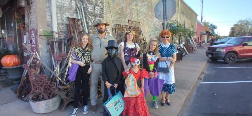 A group of people in costumes poses outside a shop, surrounded by vintage items and decorations.
