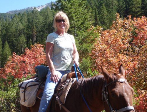 A woman smiles while riding a brown horse through a colorful forest with autumn foliage.