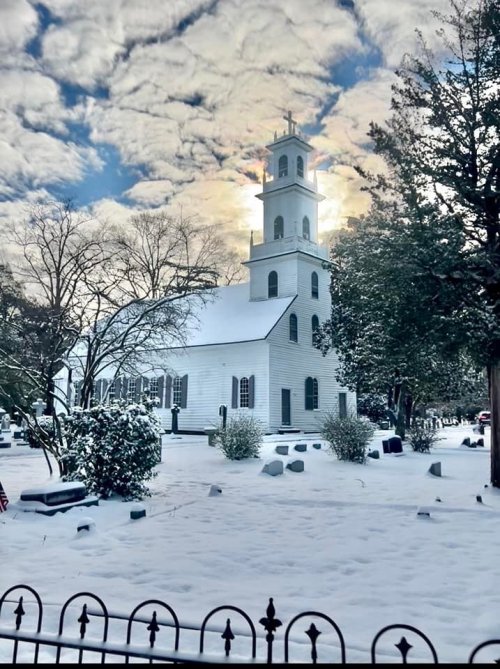 A snow-covered church with a tall steeple, surrounded by trees and gravestones under a cloudy sky.