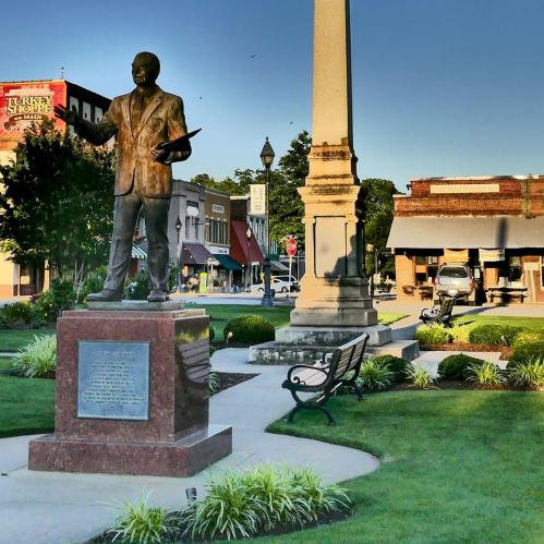 A bronze statue of a man holding a book stands in a park, surrounded by greenery and buildings in the background.