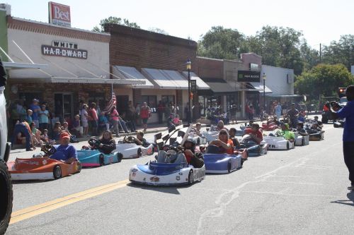A parade of children in colorful go-karts on a sunny street, with shops and spectators in the background.