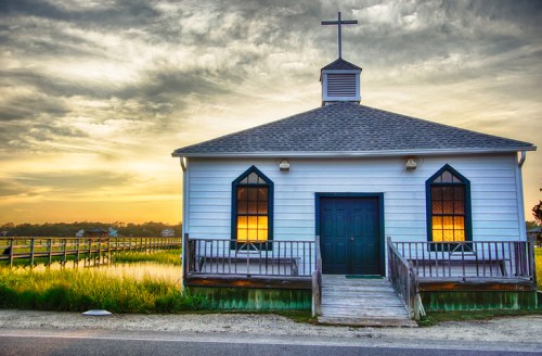 A quaint white church with a cross on top, surrounded by marshland, reflecting a sunset in its windows.