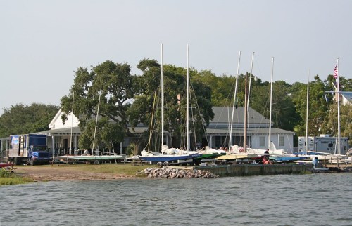 A marina with several sailboats docked near a white building surrounded by trees and water.