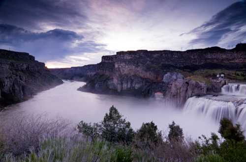 A serene landscape featuring a waterfall cascading into a river, surrounded by cliffs and a cloudy sky at dusk.