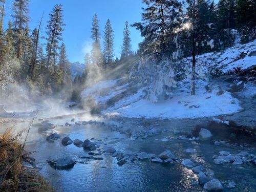 A serene winter landscape featuring a steaming river surrounded by snow-covered trees and mountains in the background.