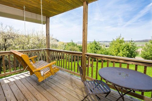 A wooden porch with a swing and a table, overlooking a green landscape and blue sky.