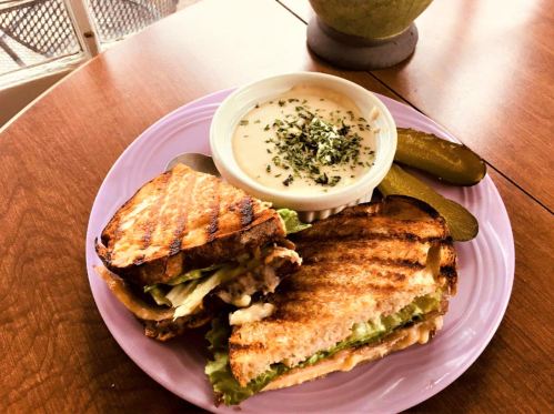A grilled sandwich with lettuce, served with a bowl of creamy soup and pickles on a purple plate.