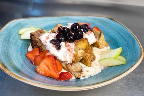 A blue plate with bread pudding topped with whipped cream, berries, and fresh fruit slices.