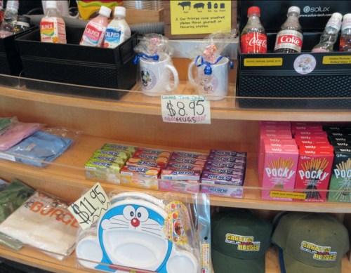 A display of various snacks, drinks, and merchandise, including mugs, plates, and hats, with price tags visible.
