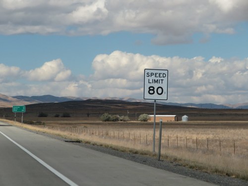 A highway scene featuring a speed limit sign reading 80, with mountains and clouds in the background.