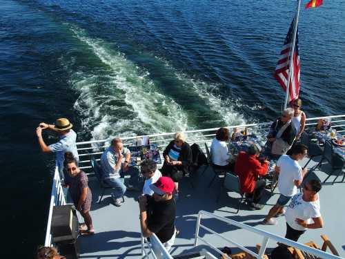 A view from a boat deck with people socializing, dining, and enjoying the water, with a flag waving in the background.