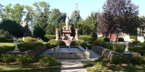 A serene garden featuring a fountain, statues, and lush greenery, with a statue at the center surrounded by flowers.