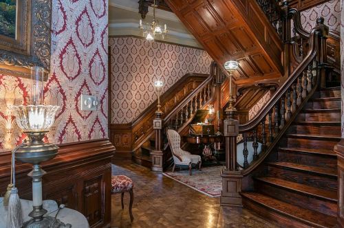 Elegant staircase in a vintage home, featuring ornate woodwork, patterned wallpaper, and a piano in the corner.
