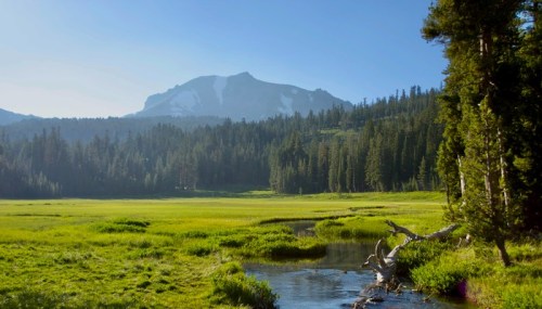 A serene landscape featuring a clear stream, lush green meadows, and mountains in the background under a blue sky.