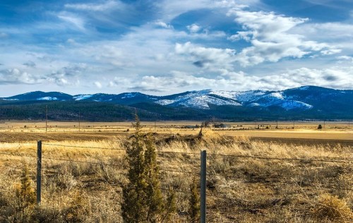 A scenic landscape featuring rolling hills, snow-capped mountains, and a clear blue sky over a golden field.