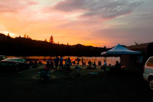 A group of people gathered by a lake at sunset, with a tent and vehicles nearby, surrounded by trees and mountains.