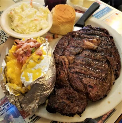 A plate featuring a large grilled steak, a baked potato with toppings, coleslaw, and a roll.