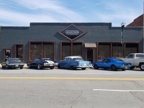A row of classic cars parked in front of Legends Steakhouse, a brick building with large windows.