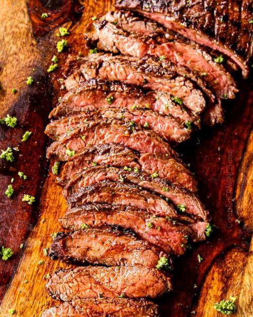 Sliced grilled steak on a wooden board, garnished with green herbs.