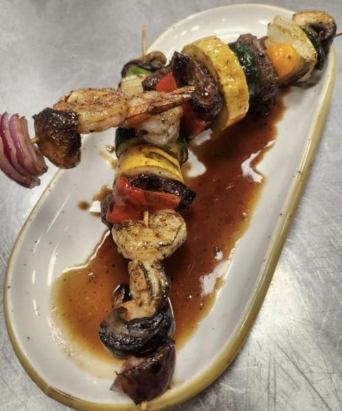 Grilled shrimp and beef skewer with colorful vegetables, drizzled with sauce, served on a white plate.