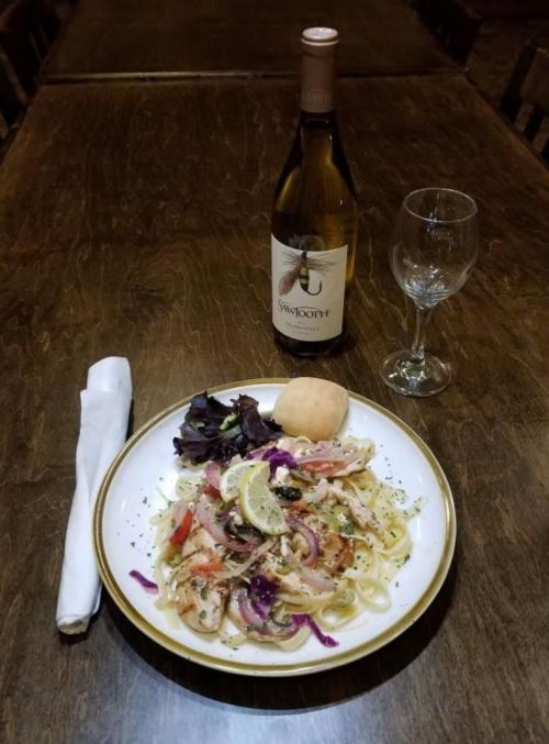A plate of seafood pasta with salad and a roll, accompanied by a bottle of white wine and a wine glass on a wooden table.