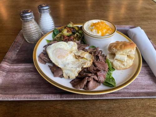 A plate of food featuring sliced beef, a fried egg, roasted vegetables, cheese-covered rice, a biscuit, and condiments.