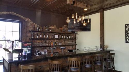 A rustic bar with a wooden counter, high stools, and a brick wall, featuring hanging lights and a TV.