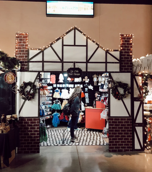 A festive shop entrance decorated with lights and wreaths, showcasing various dolls and toys inside.