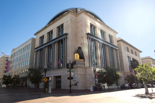 A modern building with large windows and a prominent statue on the corner, surrounded by trees and clear blue skies.
