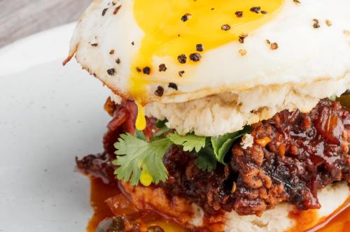 A close-up of a biscuit sandwich topped with a fried egg, cilantro, and spicy sauce, with yolk dripping down.