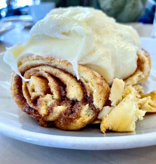 A close-up of a cinnamon roll topped with creamy frosting on a white plate.