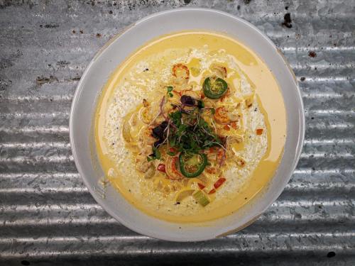 A creamy soup topped with sliced jalapeños, herbs, and crispy onions in a white bowl on a textured metal surface.