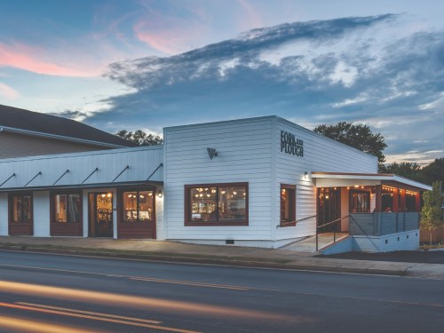 A modern white building with large windows, featuring a sign that reads "Food and Plough," set against a twilight sky.