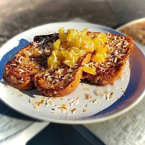 A plate of French toast topped with pineapple chunks and shredded coconut, served outdoors.