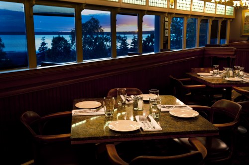 A cozy restaurant table set for dinner, with plates and glasses, overlooking a scenic view of a lake at sunset.