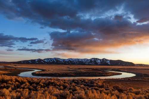 A winding river reflects the sunset, surrounded by grassy fields and snow-capped mountains under a cloudy sky.