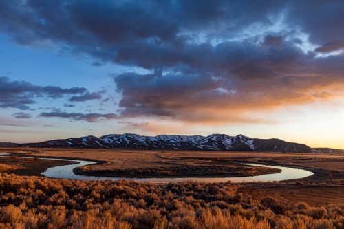 A winding river reflects the sunset, surrounded by grassy fields and snow-capped mountains under a cloudy sky.