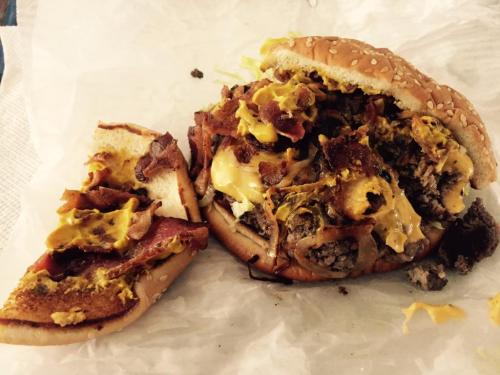 A large burger with bacon, melted cheese, and mustard, partially eaten, on a crumpled paper background.
