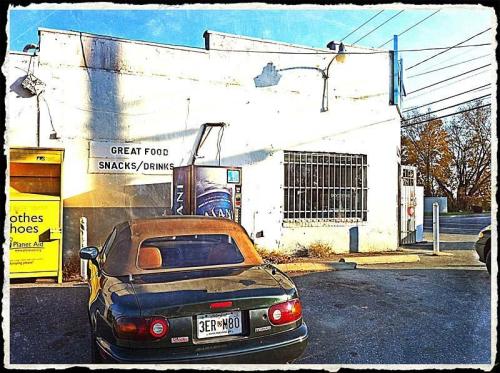 A small, weathered building with a sign for snacks and drinks, featuring a parked convertible car in the foreground.